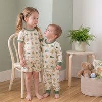 Two young children wearing matching pajamas stand together in a cozy room, with a basket of toys and a potted plant nearby.