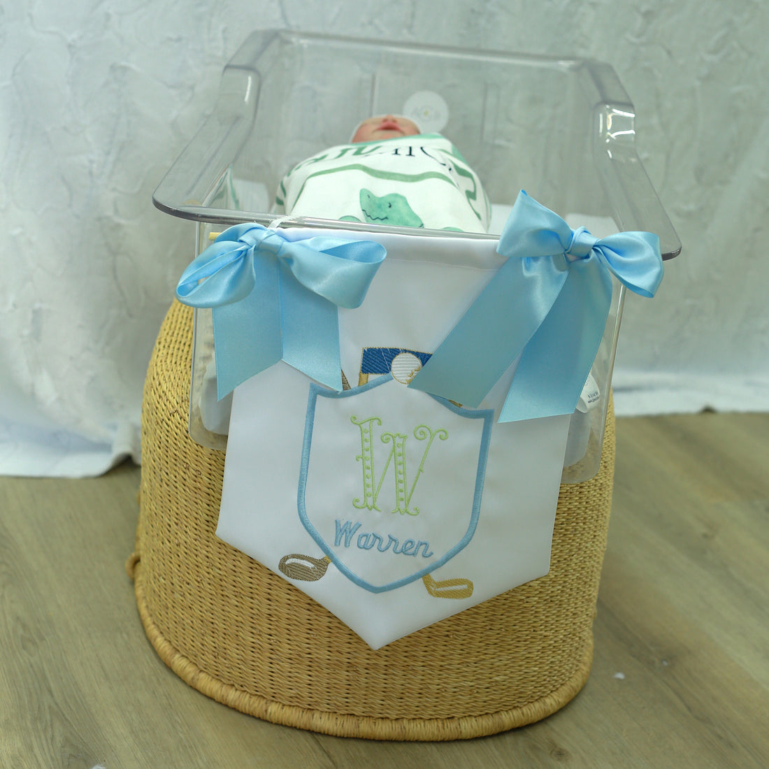 white satin door hanger for baby nursery or hospital room on the end of a newborn bassiet, blue bows on the corners, hanging string, embroidered initial and name with golf clubs and ball