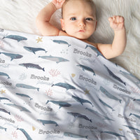 A baby is lying on a white fur-covered surface, wrapped in a blanket with a repeating pattern of blue and gray sea creatures, including sharks and starfish.