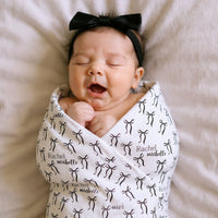 A newborn baby girl is peacefully sleeping, swaddled in a white blanket with black bows and the names "Rachael" and "Michael" printed on it.