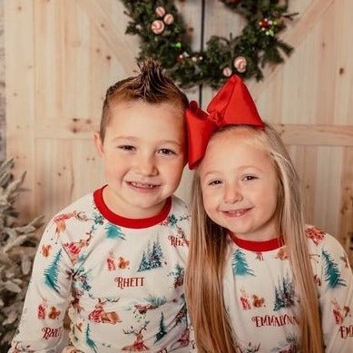 little kids wearing Christmas in the Forest soft and stretchy Christmas Pajamas with forest animals, trees, and pinecone bundles decorated for the holidays, on white fabric and red cuffs, with child's name scattered among the images.