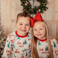little kids wearing Christmas in the Forest soft and stretchy Christmas Pajamas with forest animals, trees, and pinecone bundles decorated for the holidays, on white fabric and red cuffs, with child's name scattered among the images.