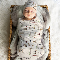 A sleeping baby swaddled in a blanket with various animal prints, including deer, moose, and bears, is lying in a woven basket.
