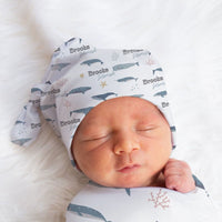 A sleeping newborn baby swaddled in a white blanket with a pattern of blue and gray sea creatures, wearing a hat with the name "Brooks" printed on it.