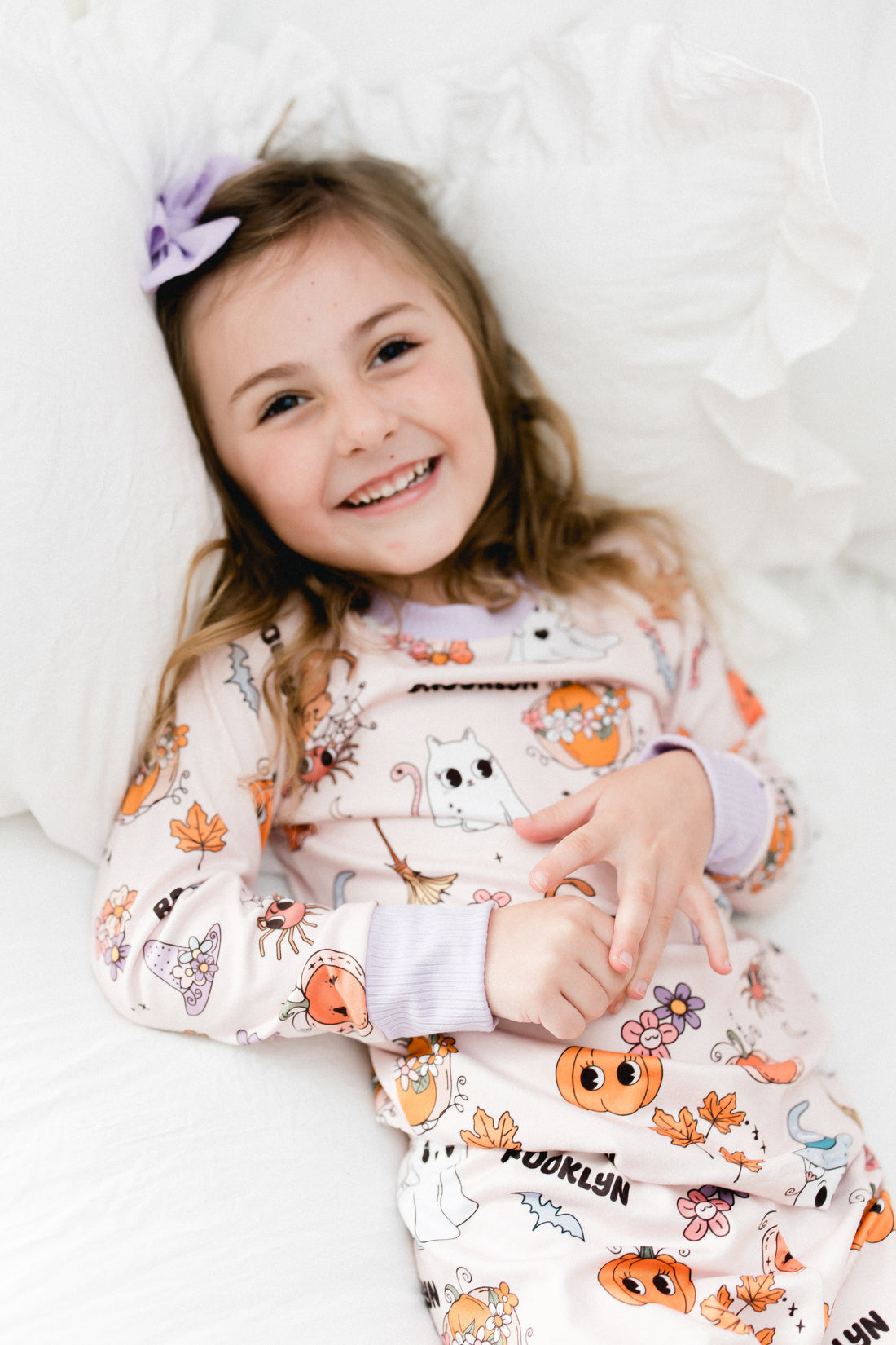 little girl wearing fall pajamas for kids that have cute pumpkins, flowers, cats and ghosts on a pale pink background and lavender cuffs.