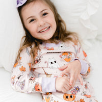 little girl wearing fall pajamas for kids that have cute pumpkins, flowers, cats and ghosts on a pale pink background and lavender cuffs.