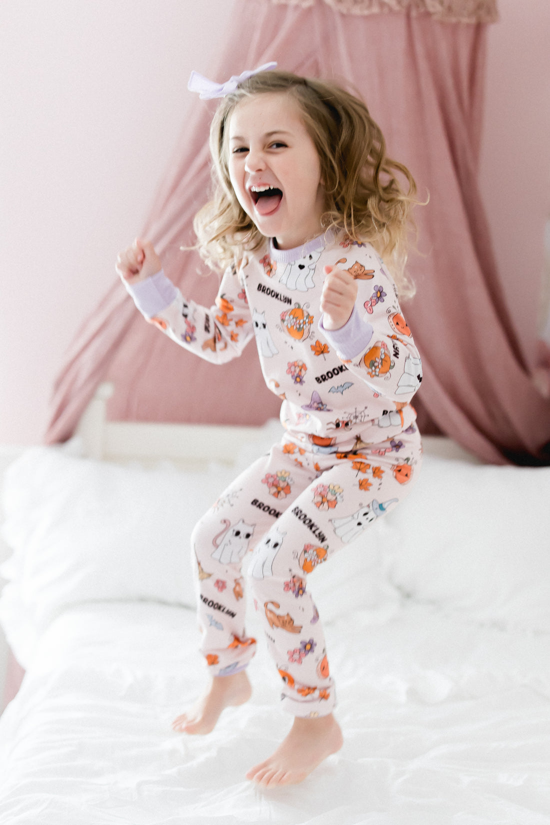 little girl wearing fall pajamas for kids that have cute pumpkins, flowers, cats and ghosts on a pale pink background and lavender cuffs.