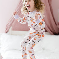 little girl wearing fall pajamas for kids that have cute pumpkins, flowers, cats and ghosts on a pale pink background and lavender cuffs.