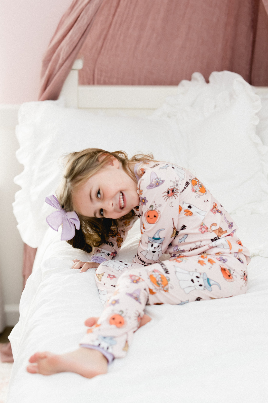 little girl wearing fall pajamas for kids that have cute pumpkins, flowers, cats and ghosts on a pale pink background and lavender cuffs.