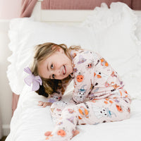 little girl wearing fall pajamas for kids that have cute pumpkins, flowers, cats and ghosts on a pale pink background and lavender cuffs.