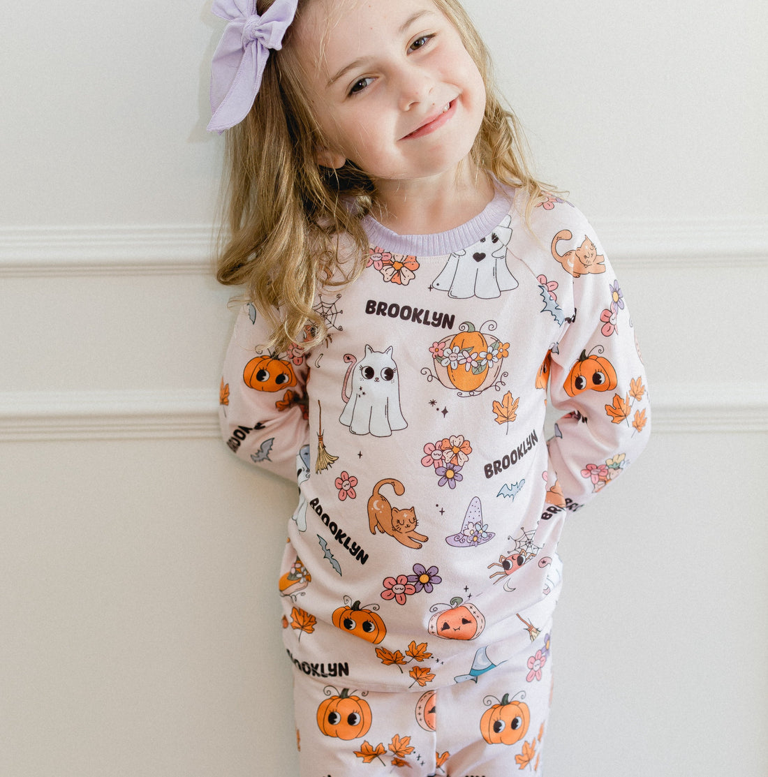 little girl wearing fall pajamas for kids that have cute pumpkins, flowers, cats and ghosts on a pale pink background and lavender cuffs.