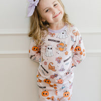 little girl wearing fall pajamas for kids that have cute pumpkins, flowers, cats and ghosts on a pale pink background and lavender cuffs.