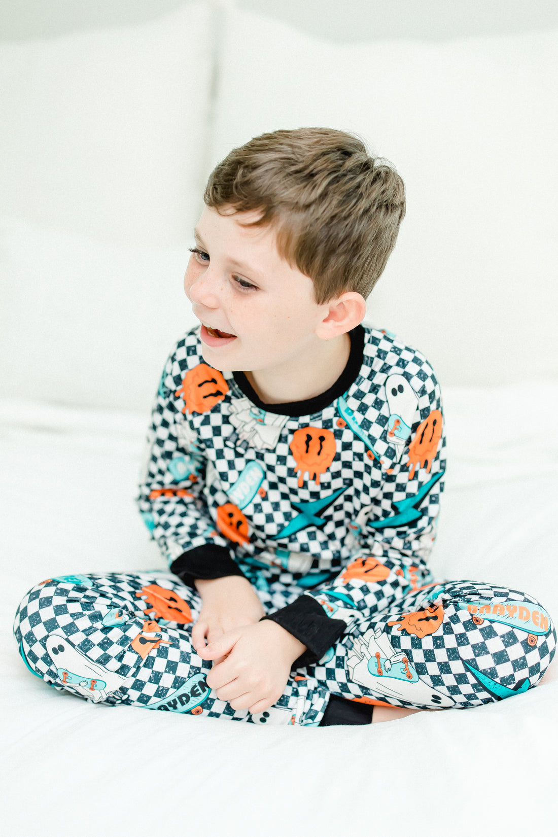 little boy wearing personalized halloween PJ's that have ghosts and skateboards and jack-o-lanterns on a black and white checkered background and black cuffs.