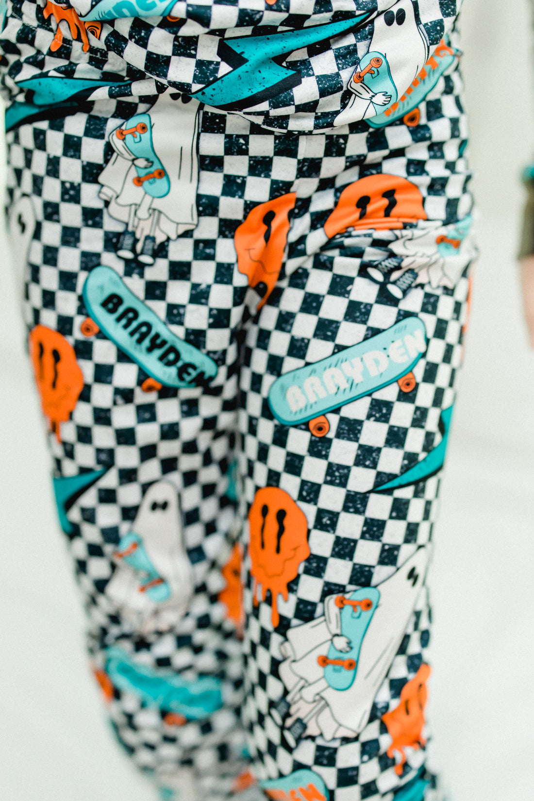 little boy wearing personalized halloween PJ's that have ghosts and skateboards and jack-o-lanterns on a black and white checkered background and black cuffs.