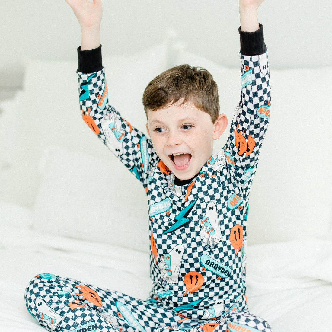 little boy wearing personalized halloween PJ's that have ghosts and skateboards and jack-o-lanterns on a black and white checkered background and black cuffs.