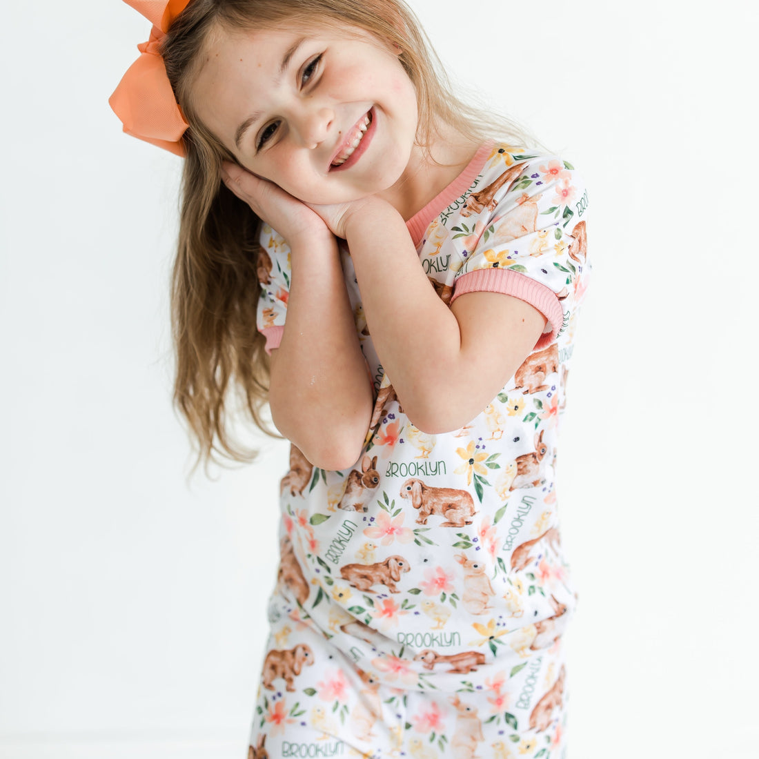 little girl wearing classic Easter bunny pajamas, Girl's custom aster pajamas with brown bunnies, and pink and yellow flowers scattered around white knit fabric along with her name, pale pink cuffs.