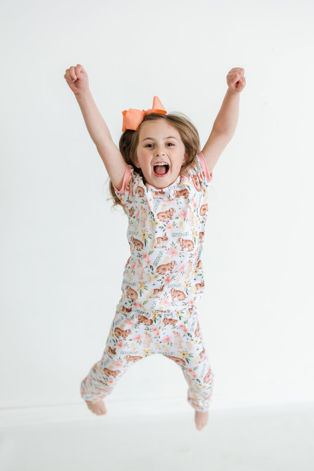 little girl wearing classic Easter bunny pajamas, Girl's custom aster pajamas with brown bunnies, and pink and yellow flowers scattered around white knit fabric along with her name, pale pink cuffs.