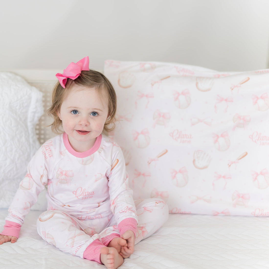 little girl laying on a personalized minky pillowcase with pink bows and baseball equipment on a pale pink background and girl's name.