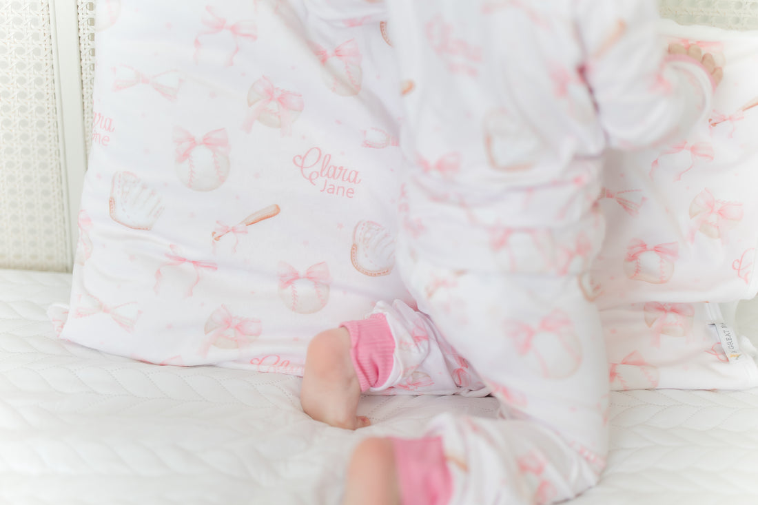 little girl laying on a personalized minky pillowcase with pink bows and baseball equipment on a pale pink background and girl's name.