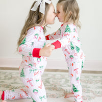 Two children in matching personalized pajamas with Christmas tree and pink stripe patterns, standing close together on a light wooden floor.