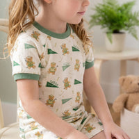 A young girl wearing a white and green pajama top with a pattern of teddy bears is sitting on a chair.