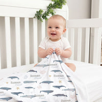 A smiling baby boy sitting in a white crib, holding a blanket with a whale and starfish pattern.