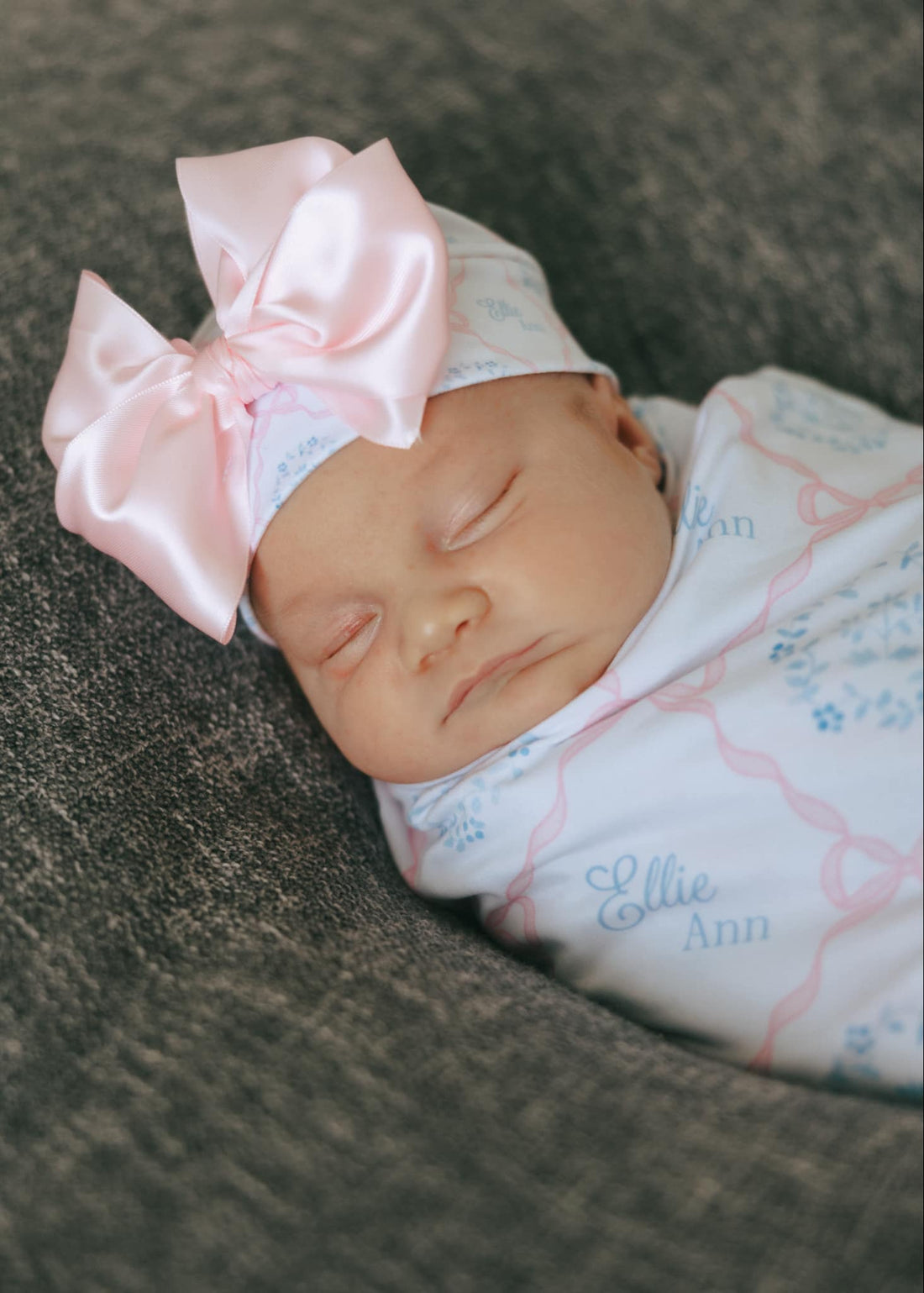 baby girl swaddled in a pink heirloom bow print personalized baby swaddle on white fabric and baby's name in light blue script font.