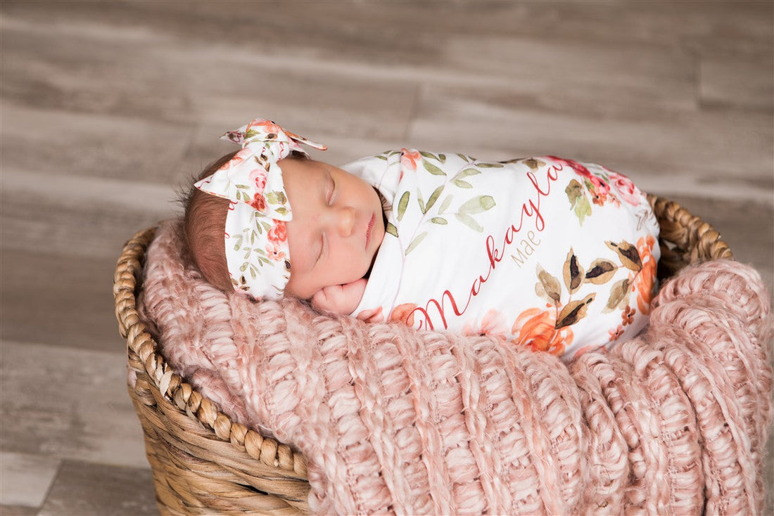 A newborn baby girl is peacefully sleeping in a basket, wrapped in a floral blanket and wearing a white headband with a bow.