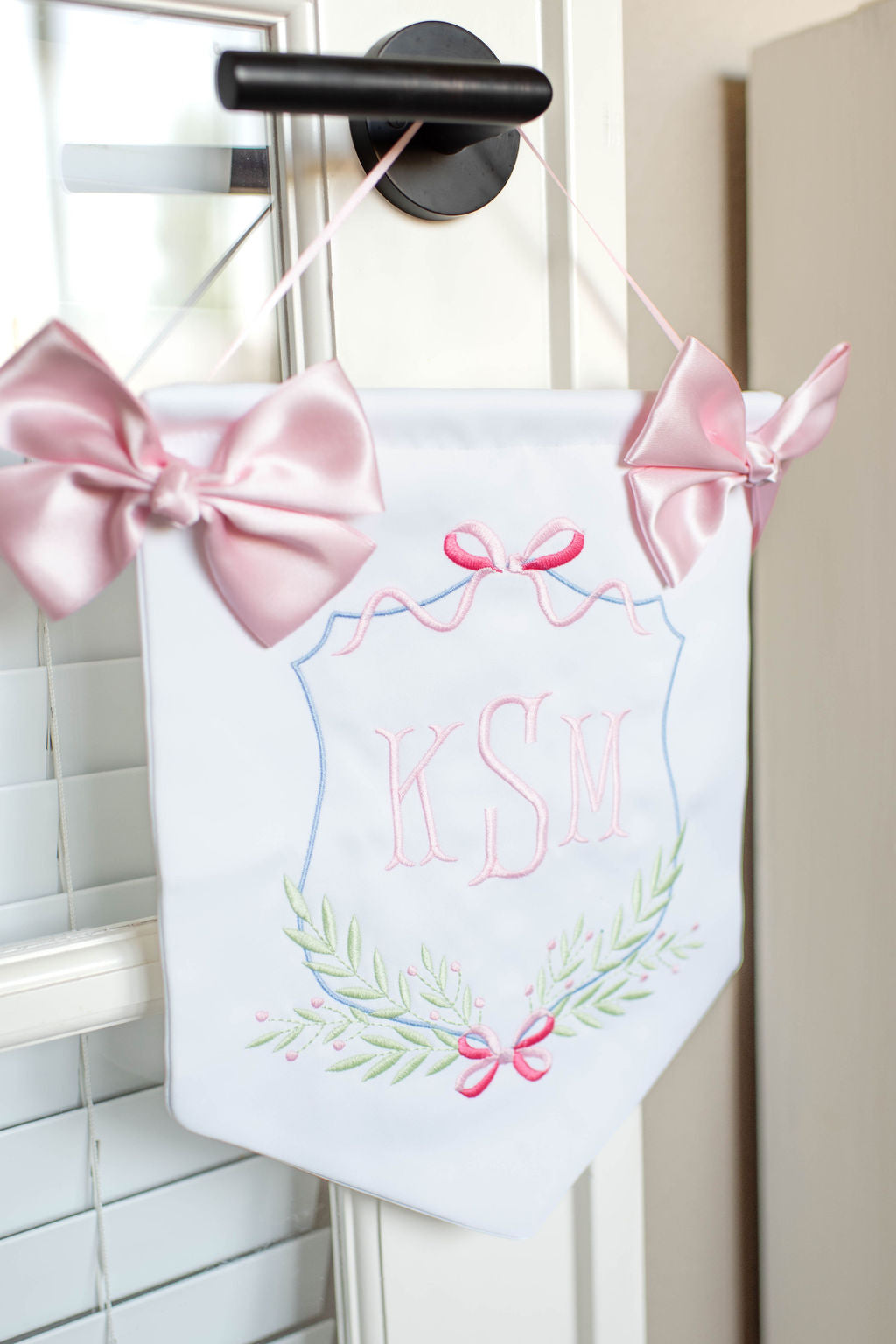 white and pink door hanging banner with baby's embroidered initials, two pink bows and a string for hanging on nursery or hospital door.