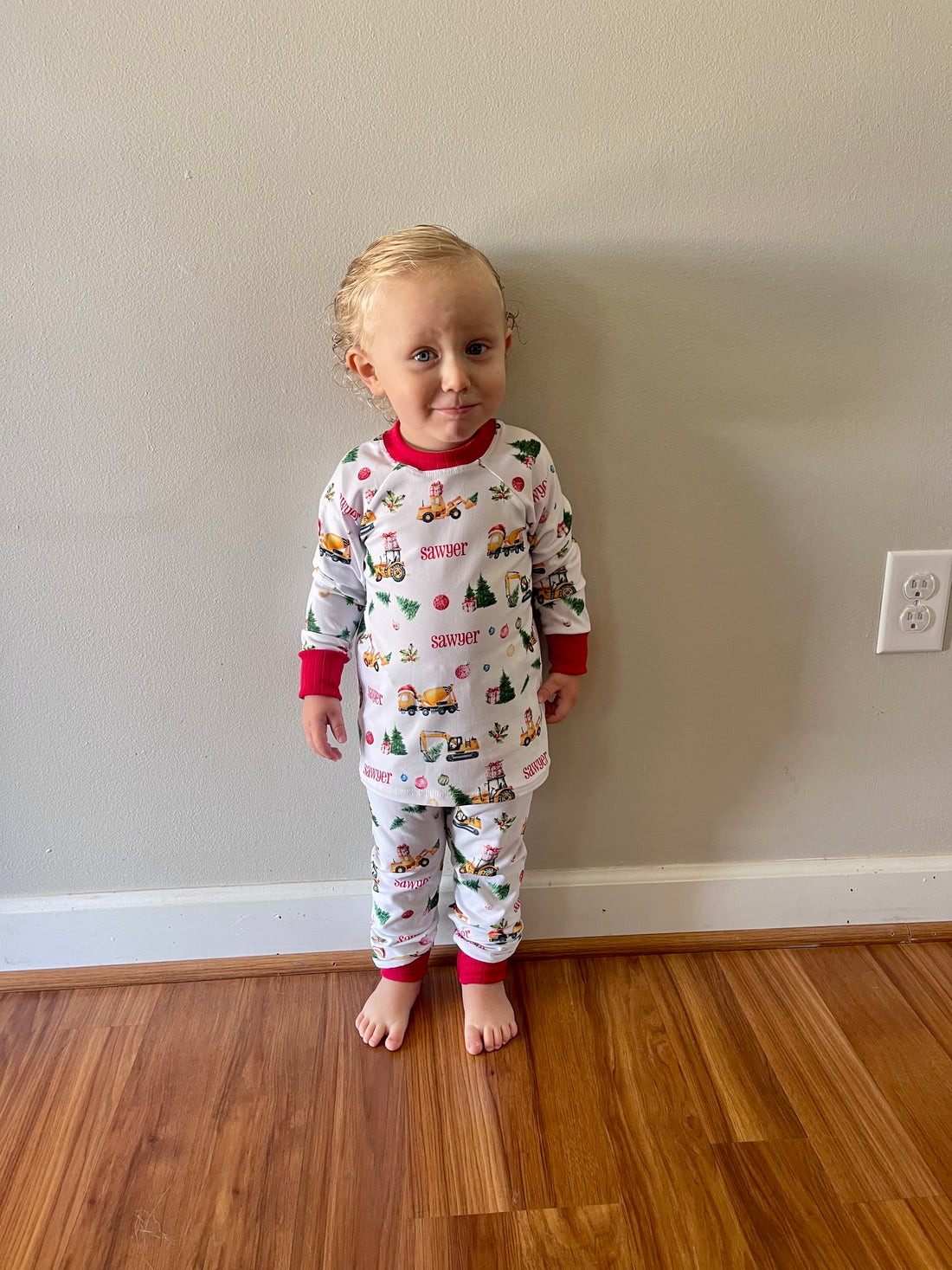 Baby standing in living room smiling while wearing Construction Christmas Pajamas.