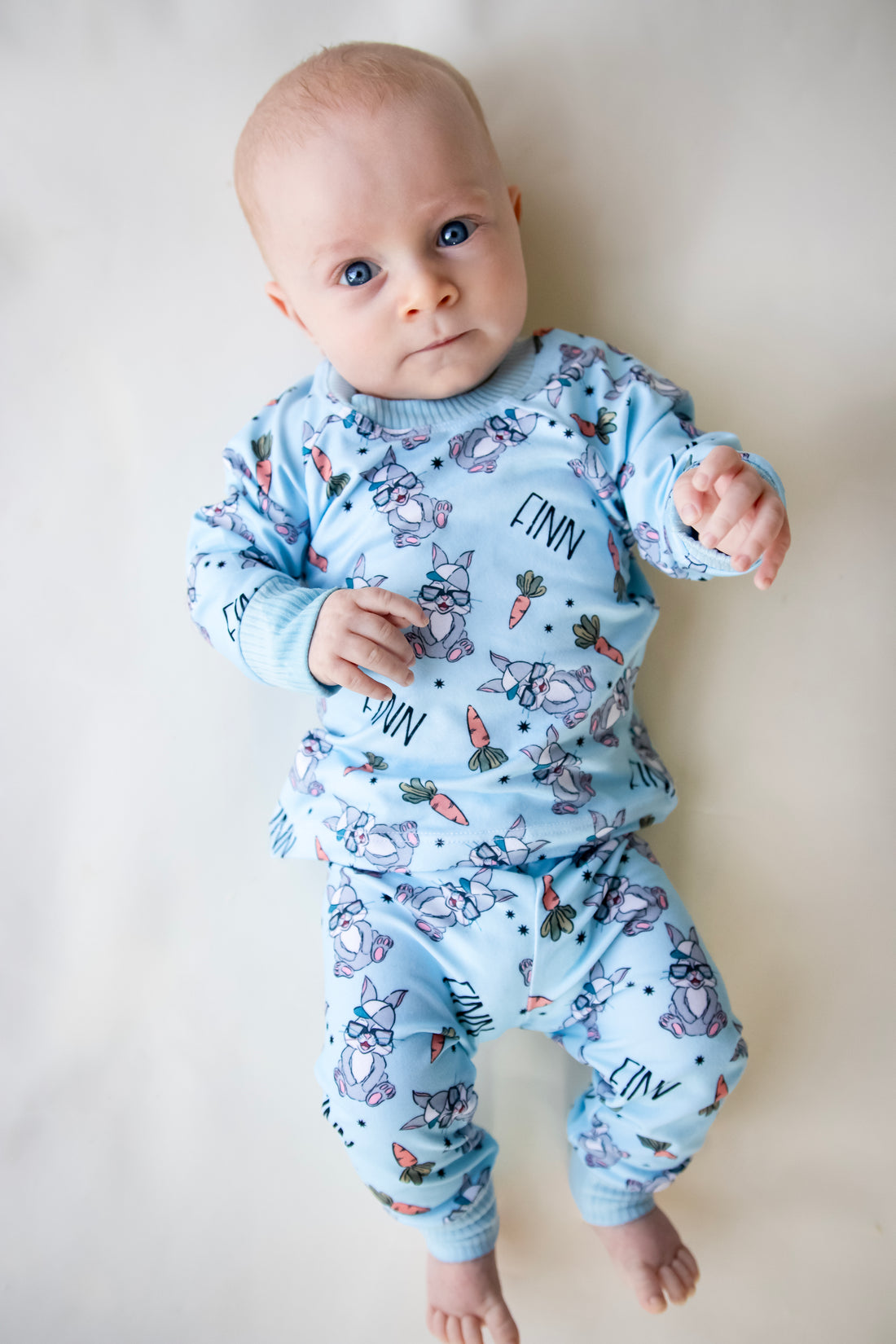 baby boy wearing boys easter personalized pajamas featuring his name, hip easter bunnies in glasses and hats, and cartoon carrots on light blue stretchy knit fabric.