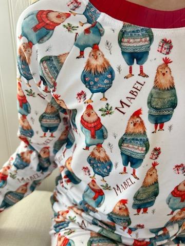 Little girl wearing personalized festive chickens christmas pajamas that feature cute chickens wearing sweaters and scarves on a white background and red cuffs.