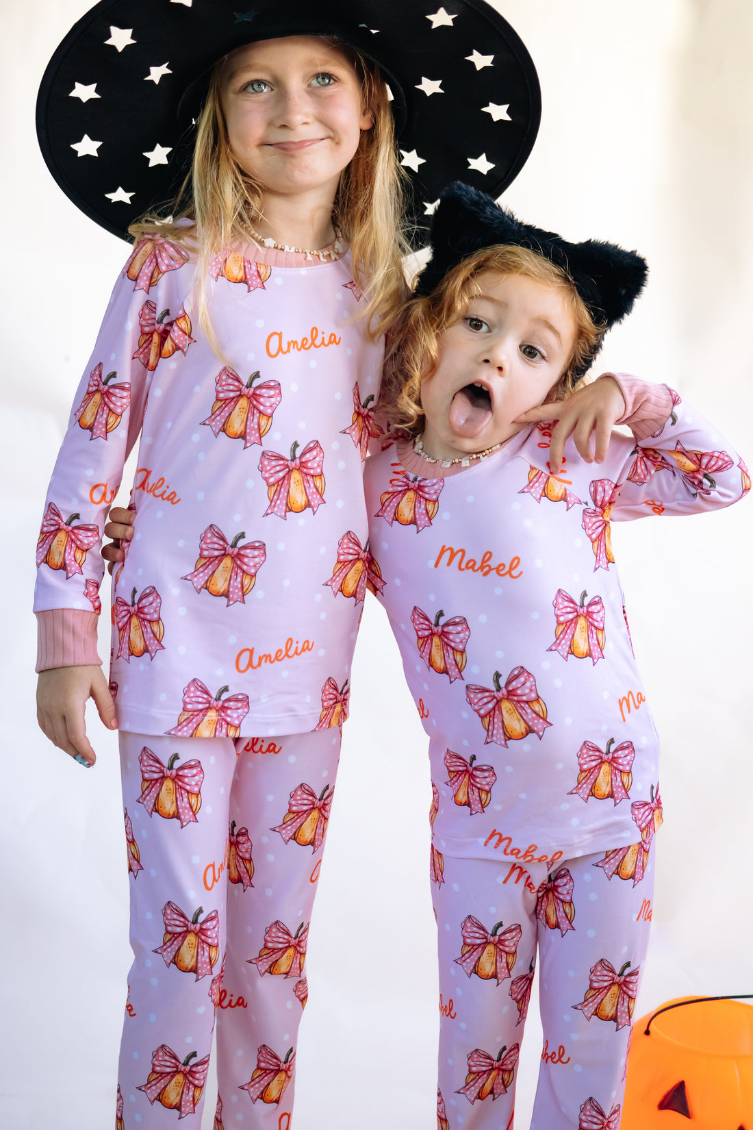 Two young girls wearing matching Halloween nightwear for kids with cartoon pumpkins and pink bows