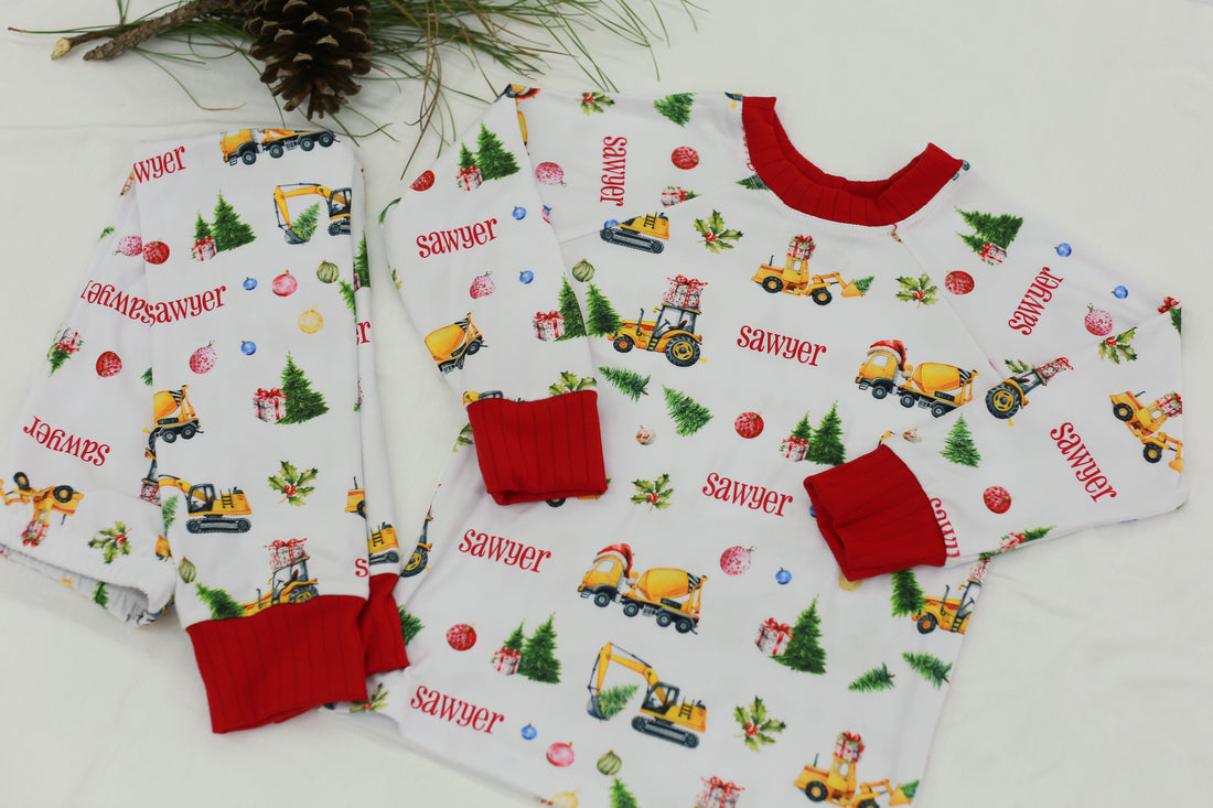 Construction Christmas Pajamas set (top and bottoms) personalized with a child's name in red font laying next to Christmas decorations.
