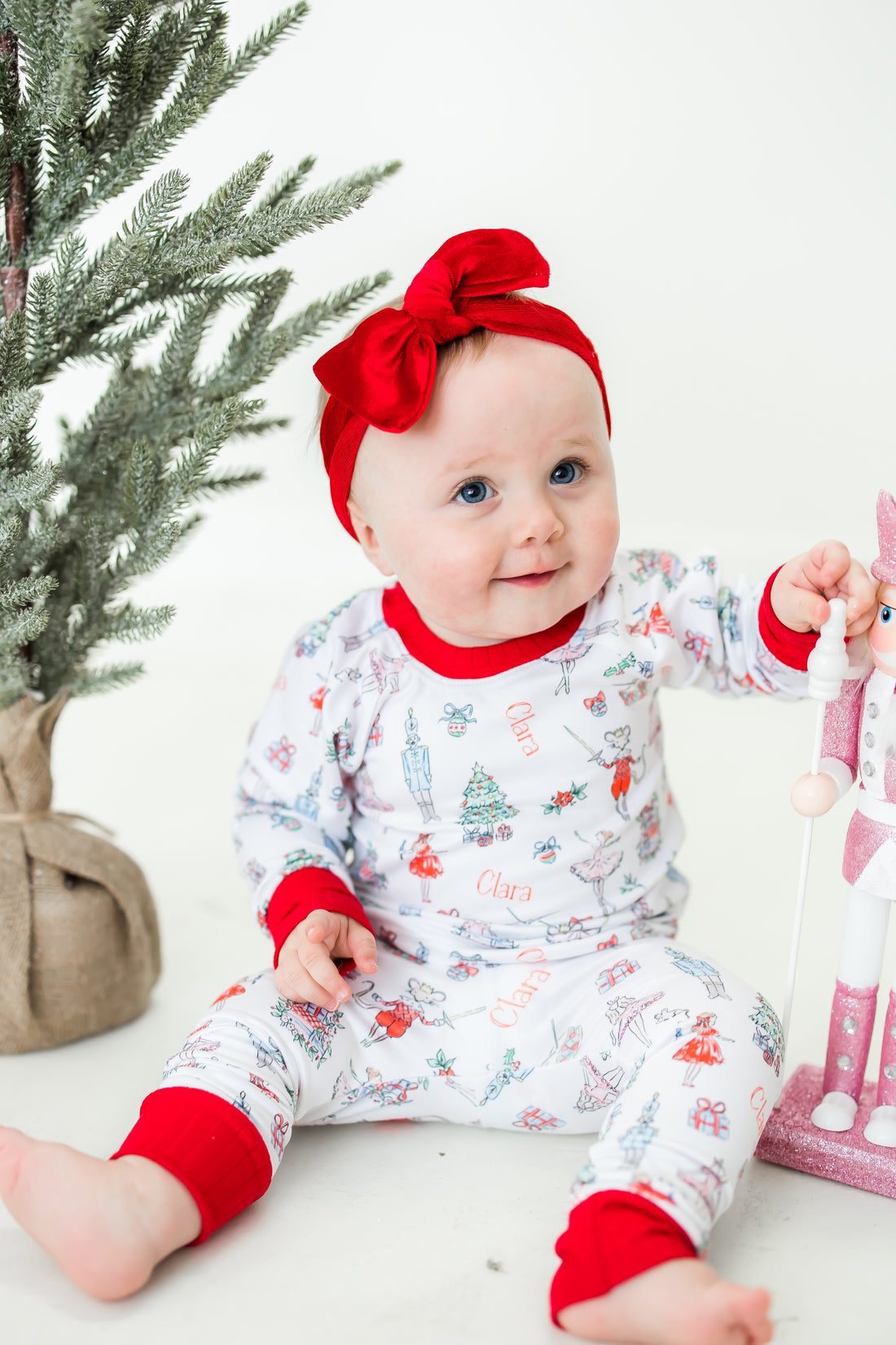 Little baby sitting by Christmas tree and wearing Nutcracker Pajamas.