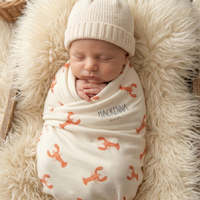 A newborn baby is peacefully sleeping, swaddled in a white blanket with an orange lobster pattern.