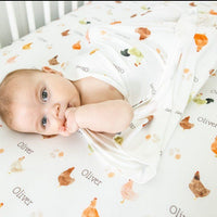 A baby is lying on a white bed with a blanket featuring a chicken pattern. The baby is looking at the camera with a curious expression.
