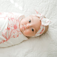 A baby girl is lying on a white bed, wearing a white floral blanket and a white headband with red flowers.