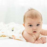 A baby lying on a white surface, looking directly at the camera with a curious expression.