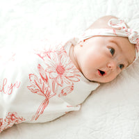 A baby girl is lying on a white bed, wearing a white and red floral blanket and a white headband with a bow.