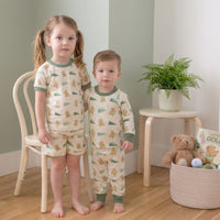 Two young children wearing pajamas with a bear pattern stand in a room with a chair, a potted plant, and a basket of toys.
