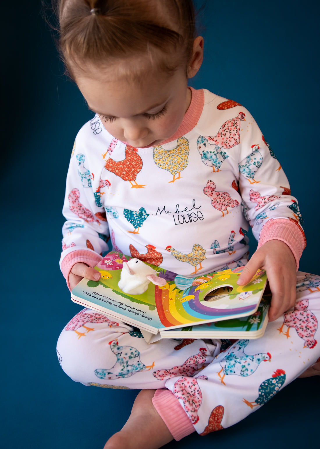 little girl wearing personalized chicken pajamas with a calico design in various colors, her name in script font on a white knit background, and pink cuffs.