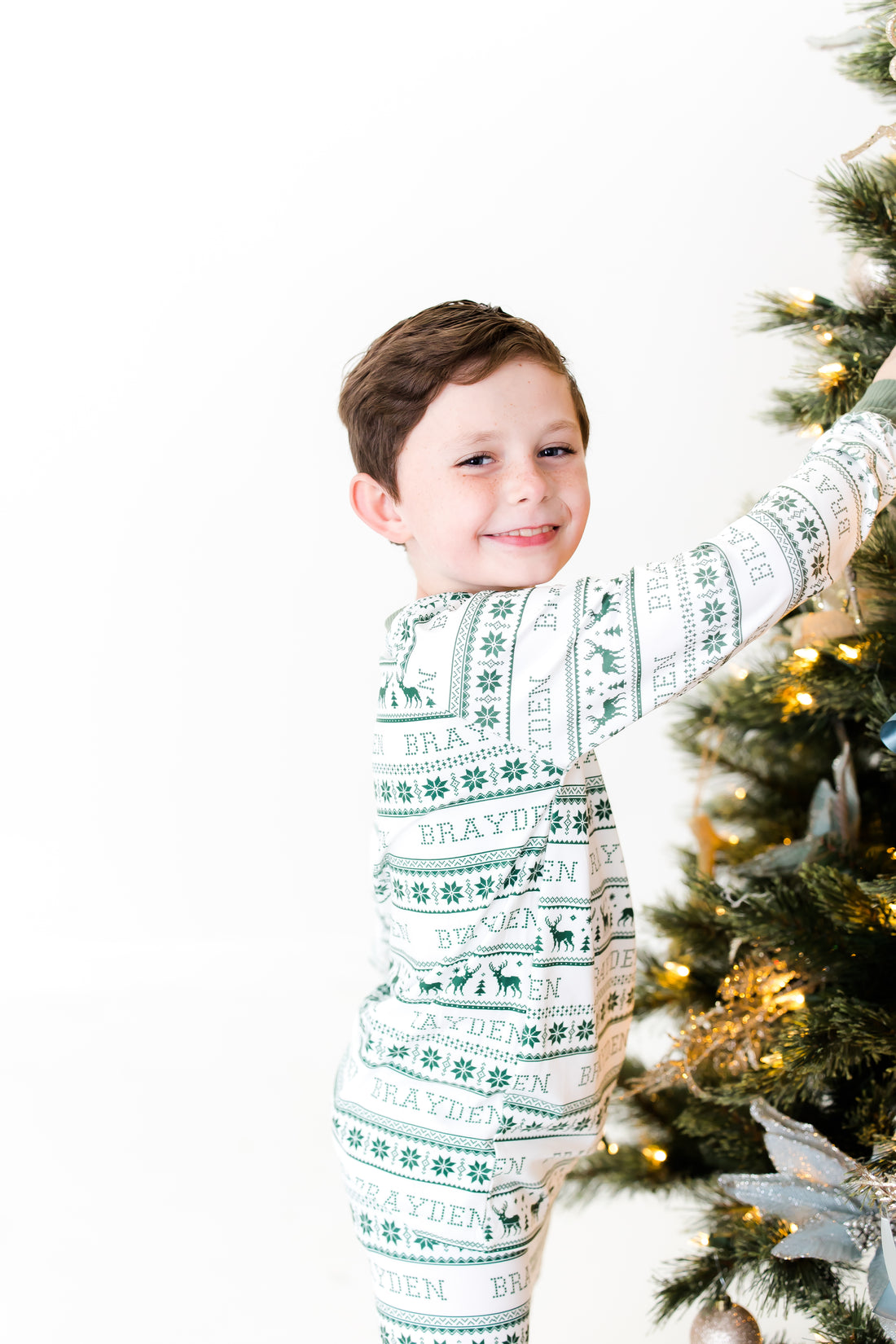 Young boy wearing green striped Christmas Pajamas and hanging ornaments on Christmas tree.