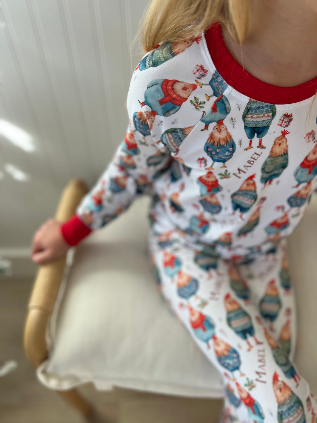 Little girl wearing personalized festive chickens christmas pajamas that feature cute chickens wearing sweaters and scarves on a white background and red cuffs.