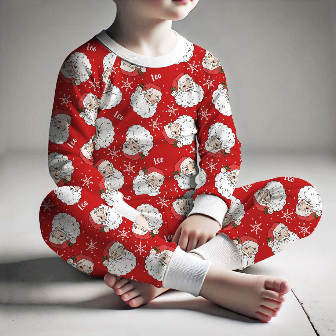 little boy wearing personalized holiday pajamas featuring vintage Santa faces and his name and snowflakes on a bright red background and white cuffs.