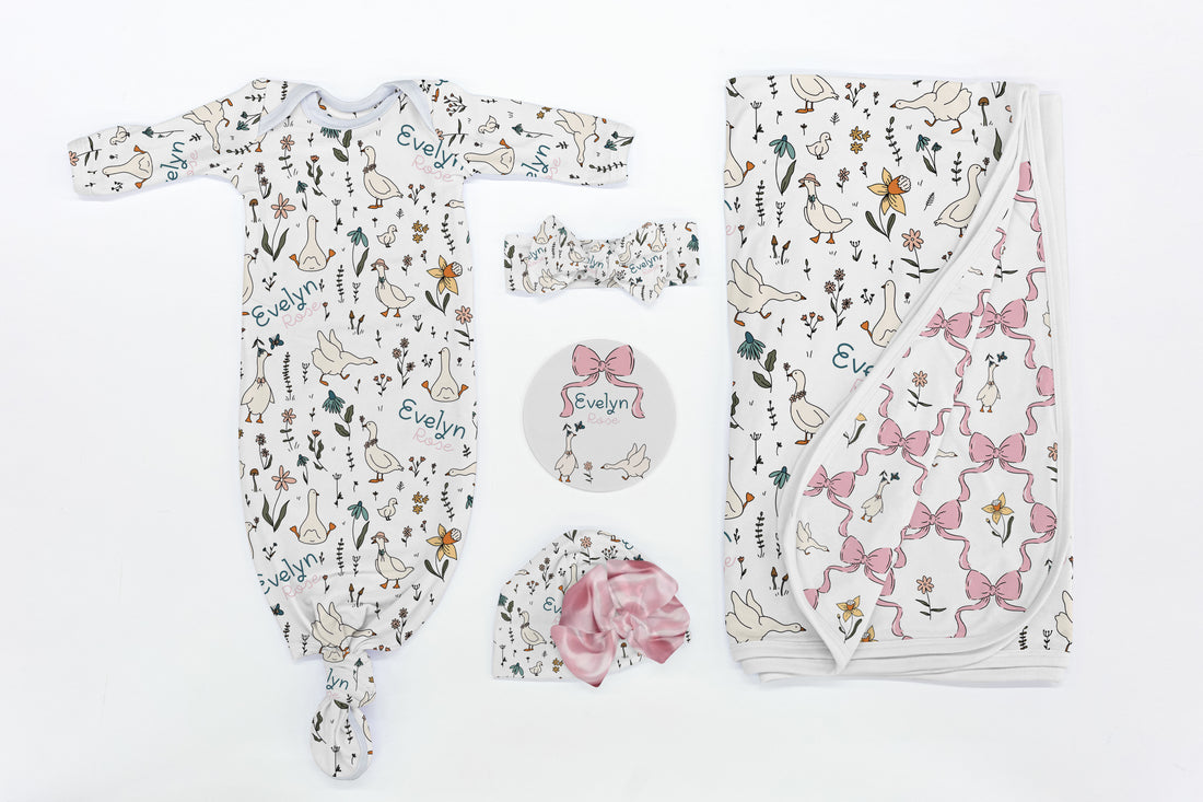 Baby clothing set with onesie, hat, and blanket featuring geese and floral and bow designs on a white background.