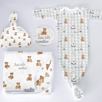 Baby clothing set with hat, blanket, and onesie featuring teddy bear pattern on a light gray background.