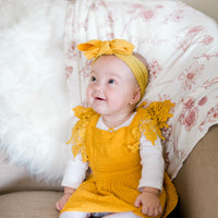 A baby girl wearing a yellow dress and a yellow headband is sitting on a couch, looking up and smiling.