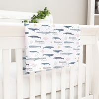 A white crib with a white blanket featuring a repeating pattern of blue and gray whale illustrations, along with the names "Roman" and "Daniel" written on it.
