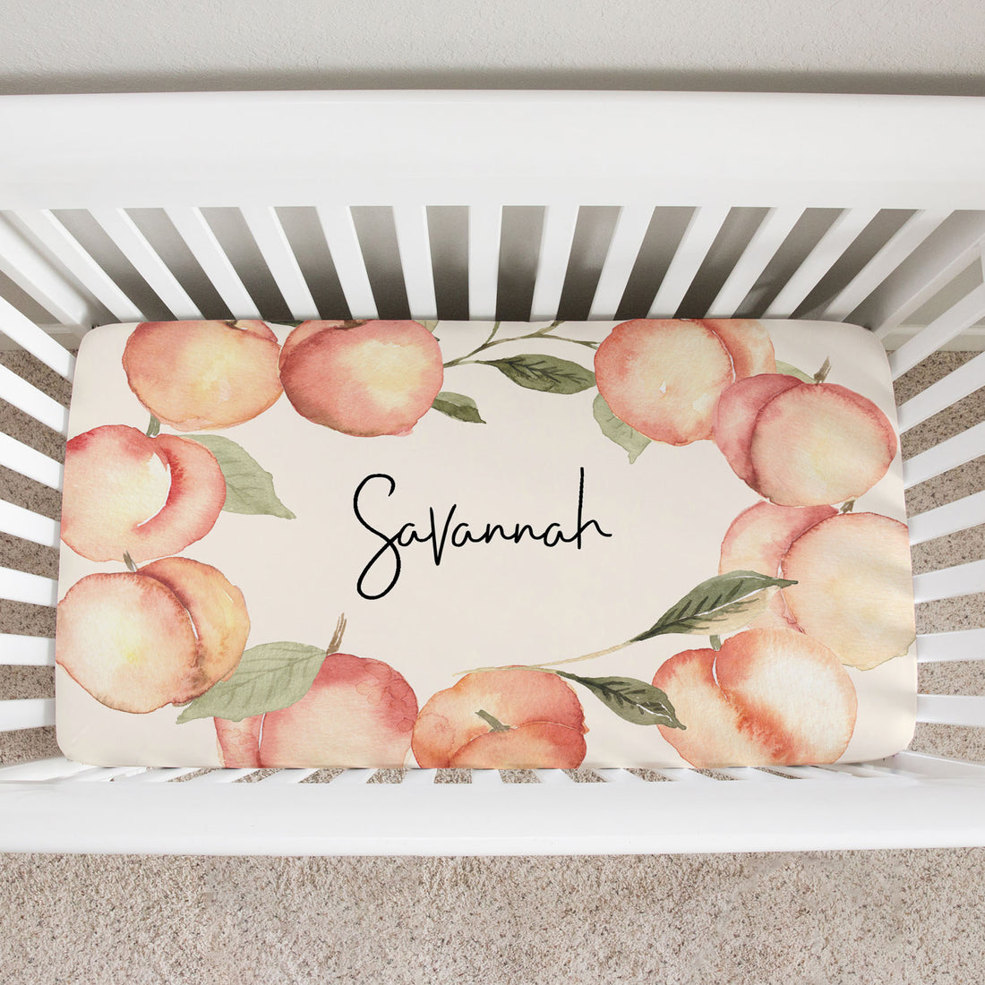 top view of a custom crib sheet. It has a frame of big watercolor peaches for the Georgia Peach in your life. Her name is written in the middle with a modern font.